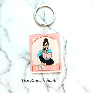 May include: A pink and white keychain with a cartoon illustration of a person wearing glasses and reading a book while holding a cup of coffee. The text "The Reader" is printed below the illustration. The text "The Rensch Nook" is printed below the keychain.