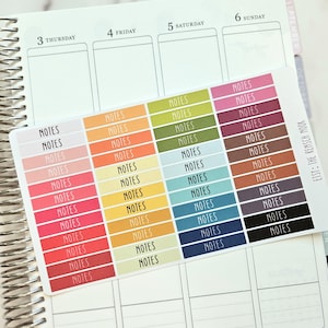 May include: A planner with a spiral binding, open to a weekly view. A sticker sheet with the word "NOTES" in various colors is placed on the planner. The planner has the days of the week printed at the top.