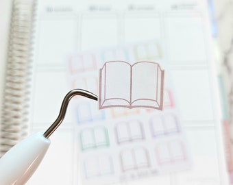 May include: A clear sticker sheet with multiple book icons. The icons are in various colors, including pink, blue, green, and yellow. The icons are outlined in black. The sticker sheet is on a white surface.