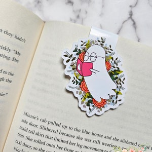 May include: A white ghost with glasses reading a pink book surrounded by floral details. The bookmark is a great gift for book lovers.