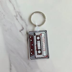 May include: A rectangular keychain featuring a cassette tape design. The tape is gray with red accents and the word "THRILLER" in white. A silver key ring is attached to the top.