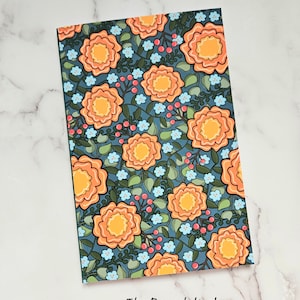 May include: A rectangular notebook with a floral pattern. The design features large orange flowers with yellow centers, small blue flowers, and red berries on a dark teal background. The text "The Rensch Nook" is at the bottom.