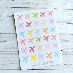 May include: A white sticker sheet featuring a grid of small airplane icons in various pastel colors including pink, yellow, blue, green, and orange. The bottom of the sheet displays the black printed text: ETSY: THE RENSCH NOOK.
