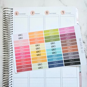 Puede incluir: Una hoja de pegatinas coloridas con la palabra "GOALS" impresa en cada pegatina. Las pegatinas est&aacute;n dispuestas en filas y columnas, con cada fila con un color diferente. Las pegatinas est&aacute;n dise&ntilde;adas para usar en una agenda o diario.