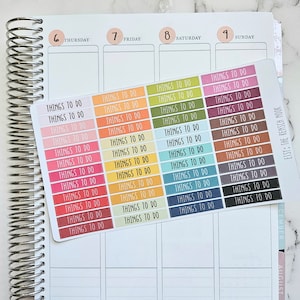 May include: A planner is open to a double-page spread, with a sticker sheet of colorful "Things To Do" labels. The planner has a silver spiral binding. The sticker sheet includes labels in shades of pink, orange, green, blue, and brown.