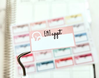 Puede incluir: Una pegatina rosa y blanca con el texto "ENT appt" en un planificador blanco con un gancho plateado.