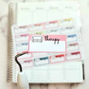 May include: A pink sticker with the word "therapy" in cursive and a sofa icon. The sticker is held by a white and silver tool, with a planner and other stickers in the background. The stickers are for planning and organization.