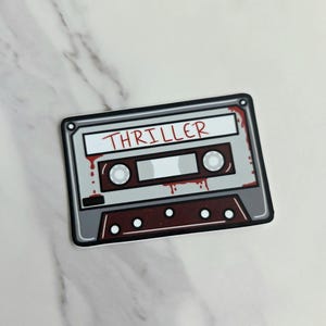 May include: A sticker featuring a cassette tape design with the word "THRILLER" in red. The cassette tape is gray and brown with red blood splatter details. The sticker is on a white and gray marble surface.