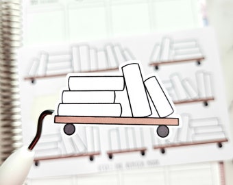 May include: A sticker featuring a stack of white books on a brown cart with black wheels. The sticker is placed on a planner with a blurred background of bookshelves. The cart has a black handle.