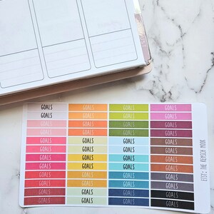 Puede incluir: Una hoja de pegatinas de colores con la palabra "GOALS" impresa en cada pegatina. Las pegatinas est&aacute;n dispuestas en filas de cuatro, con cada fila presentando un color diferente. Los colores incluyen rosa, naranja, amarillo, verde, marr&oacute;n, rojo, azul y gris. Las pegatinas est&aacute;n dise&ntilde;adas para su uso en una agenda o diario.
