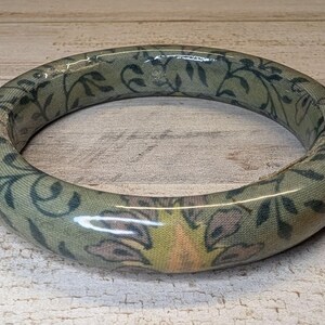 May include: A circular bangle bracelet featuring a smooth, glossy finish over a sage green fabric base. The surface is decorated with a repeating botanical pattern in dark green, muted brown, and soft yellow tones, creating a continuous floral motif.