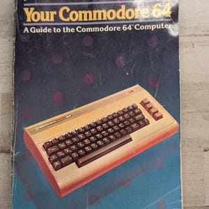 May include: A softcover book titled Your Commodore 64: A Guide to the Commodore 64 Computer by John Heilborn and Ran Talbott. The dark blue cover features a graphic illustration of a beige Commodore 64 computer with a black keyboard.