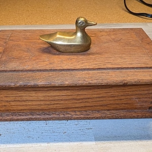 May include: A rectangular wooden box with a medium brown wood grain finish and visible finger joints at the corners. A polished brass duck figurine is mounted on the center of the lid, serving as a handle for the box. The lid features a raised panel.