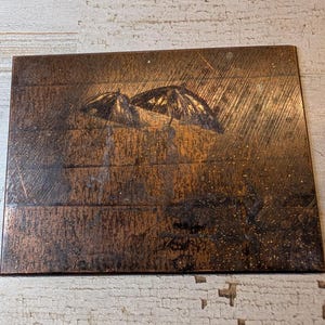 May include: A rectangular copper-toned metal plate featuring an etched design of two figures walking under umbrellas in the rain. The surface has a brushed, metallic texture with dark, weathered accents that highlight the scene of the figures.