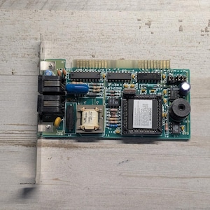 May include: A green printed circuit board featuring a metallic expansion slot bracket, two black ports, a central transformer, various resistors, capacitors, and a large integrated circuit chip with a white label containing technical text.