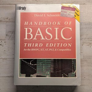 May include: A softcover book titled Handbook of BASIC Third Edition by David I. Schneider for the IBM PC, XT, AT, PS/2, & Compatibles. The cover is light gray with a large red rectangle containing white text and a black graphic of computer code.