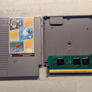 May include: A gray Nintendo Entertainment System game cartridge featuring a label with Super Mario Bros., Duck Hunt, and World Class Track Meet. The cartridge is disassembled, showing the gray plastic casing and the internal green circuit board.
