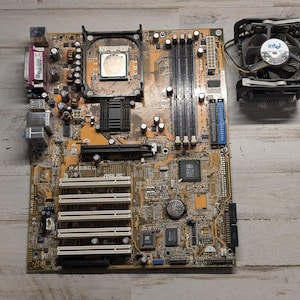 May include: An ASUS P4S800 motherboard with a tan printed circuit board, featuring a central CPU socket, four memory slots, multiple white PCI expansion slots, and various integrated components, accompanied by a separate black Intel CPU cooler.
