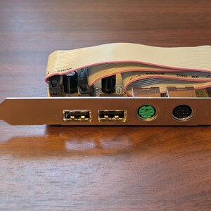 May include: A computer expansion card featuring a metal bracket with two USB ports, one green PS/2 port, and one black circular port. A beige ribbon cable is connected to the circuit board, which includes black capacitors and electronic components.