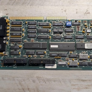 May include: A green printed circuit board featuring a gold-plated edge connector, two black external ports on a metal bracket, and numerous integrated circuits, resistors, and capacitors soldered across the surface with various white text labels.