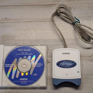 May include: A Brother PED-Basic embroidery software CD in a clear plastic case and a white and blue card reader device with an attached USB cable. The CD label features the text PED-Basic Ver 1.0 for Windows 98/ME/XP/2000 and Brother branding.