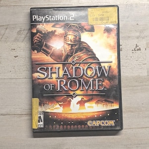 May include: A PlayStation 2 video game case for Shadow of Rome by Capcom. The cover art features a Roman soldier in armor holding a goblet, with silhouettes of gladiators in an arena below. A yellow pre-owned sticker is on the top right corner.
