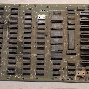 May include: A green Apple II main logic printed circuit board featuring numerous black integrated circuit chips, multiple expansion slots on the right side, and various electronic components. The board is labeled with Apple II MAIN LOGIC.