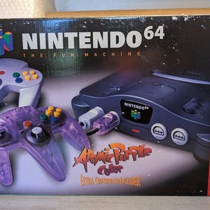 May include: A Nintendo 64 video game console box featuring a dark gray console, a solid light purple controller, and a translucent atomic purple controller. The packaging displays the Nintendo 64 logo, text, and a red side panel with numbered features.