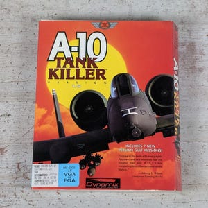 May include: A rectangular cardboard box for the A-10 Tank Killer Version 1.5 computer game. The front features a dark gray A-10 aircraft against a bright yellow sun and orange sky. Text includes the title, version, and system requirements.