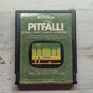 May include: A black rectangular Atari video game cartridge for Pitfall! featuring a green label with the title, game credits, and a central graphic showing a jungle scene with a character swinging on a vine over a pit and a scorpion.