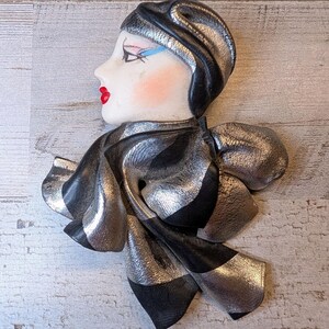 May include: A decorative wall plaque featuring a stylized face in profile, wearing a black and metallic silver headpiece and a matching scarf. The face has pale skin, red lips, and blue eyeliner. The scarf is draped in flowing, layered folds.