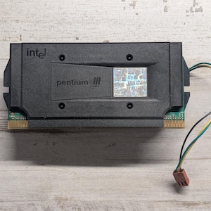 May include: An Intel Pentium III processor cartridge featuring a black plastic housing with the Intel logo and Pentium III text printed on the front. A holographic square sticker is visible on the right side, and a thin multi-colored wire with a connector.