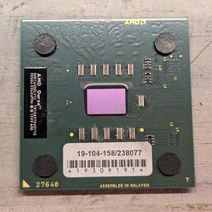 May include: A square green AMD Duron computer processor featuring a central purple die, silver surface-mounted components, and a black label with white text. A white sticker displays the serial number 19-104-158/238077 and a barcode.
