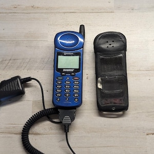 May include: A blue Qualcomm US West mobile phone with a numeric keypad, a small screen, and an antenna, connected to a black coiled car charger cable with a cigarette lighter adapter, alongside a black leather protective carrying case.