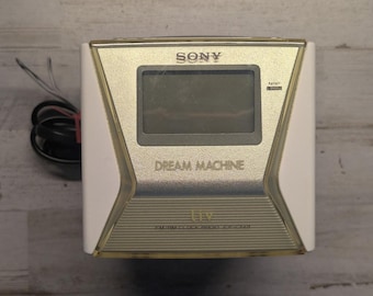 May include: A white and metallic gold Sony Dream Machine FM/AM clock radio with a rectangular digital display screen, a NAP button, and a black power cord extending from the left side. The front panel features the text SONY, DREAM MACHINE, and FM/AM CLOCK RADIO ICF-C113.