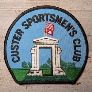 May include: A circular embroidered patch with a light blue background, a black border, and the text CUSTER SPORTSMEN'S CLUB in black. The design features a white stone archway, a green grassy field, and a small red and white flag on a pole.