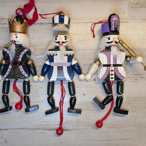 May include: Three wooden nutcracker ornaments with pull-string mechanisms. The left figure wears a gold crown and black jacket. The center figure has a blue and white crown and blue jacket. The right figure wears a purple hat and holds a gold baton.
