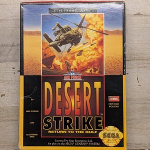 May include: A rectangular video game box for Desert Strike: Return to the Gulf for the Sega Genesis system. The cover features a military helicopter flying over a desert landscape with an explosion, the Electronic Arts logo, and a Sega seal.