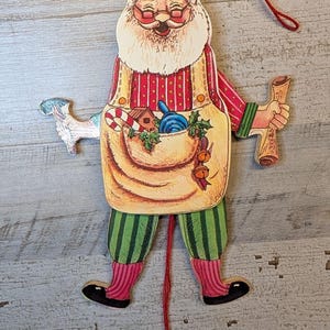 May include: A flat wooden Santa Claus ornament with a red string for hanging and a pull string at the bottom. Santa wears a tan apron over a red striped shirt and green striped pants, holding a scroll and a toy, with a candy cane in the apron pocket.