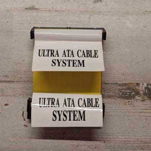 May include: A yellow ribbon cable with black connectors at both ends. Two white labels are attached to the cable, each featuring the black printed text ULTRA ATA CABLE SYSTEM. The cable has a flat, multi-wire design with a distinct ribbed texture.