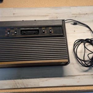 May include: A black Atari Video Computer System console featuring a wood-grain front panel, horizontal ribbed texture, and a top control panel with switches for power, TV type, game select, and reset, accompanied by a black wired power cable.
