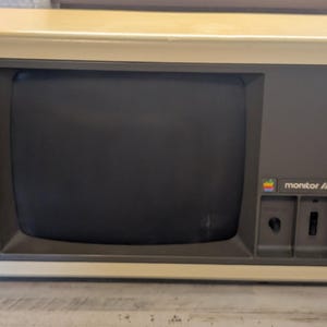 May include: A vintage beige computer monitor with a dark gray screen bezel. The front panel features the text monitor /// and a small rainbow-colored Apple logo. Below the text are two vertical control knobs for adjusting display settings.