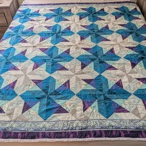May include: A rectangular quilt featuring a repeating pinwheel pattern in shades of teal, dark blue, and purple, interspersed with light gray and floral-patterned fabric blocks. The quilt is finished with a decorative border in purple and gray.