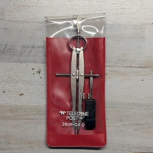 May include: A silver metal drafting compass with a black handle attachment, housed in a clear and red vinyl protective sleeve. The sleeve features the white printed text Teledyne Post 38JP-040. The tool includes a central adjustment wheel.