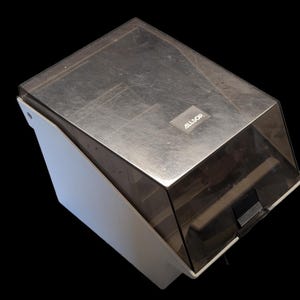 May include: A white plastic storage case with a slanted, transparent dark gray lid. The lid features a small rectangular label with the text ALLSOP printed in white. The container has a rectangular base and a hinged top cover with a black latch.