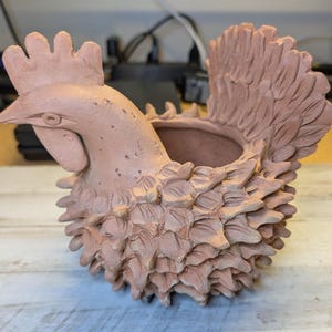 May include: A terracotta-colored ceramic planter shaped like a rooster. The vessel features a textured body with sculpted feather-like details, a prominent comb on the head, and a large, fanned tail. The center of the rooster is hollowed out.