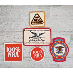 May include: Five embroidered patches featuring National Rifle Association branding. Includes a triangular 1982 patch, a rectangular patch with an eagle graphic, two red patches with 100% NRA text, and a circular red, white, and blue endowment member patch.