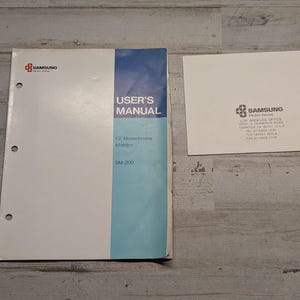 May include: A white and blue Samsung user's manual for a 12" monochrome monitor model SM-200 with three binder holes, alongside a white Samsung business card featuring the Los Angeles office address, phone, telex, and fax numbers.