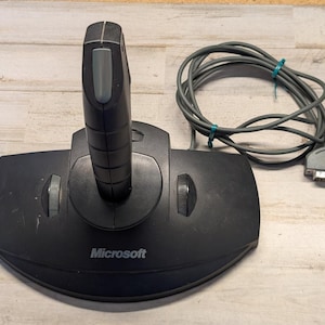 May include: A black Microsoft joystick with a vertical handle, a gray trigger button, and two side-mounted thumb wheels on the base. The device includes a long gray cable with a multi-pin connector attached to the rear of the controller base.