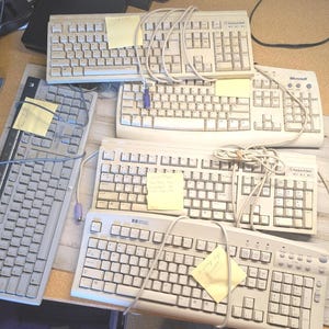 May include: A collection of five beige and grey computer keyboards with QWERTY layouts, each featuring a coiled or straight cable and a small yellow adhesive note attached to the surface. The keyboards vary in brand and key arrangement.